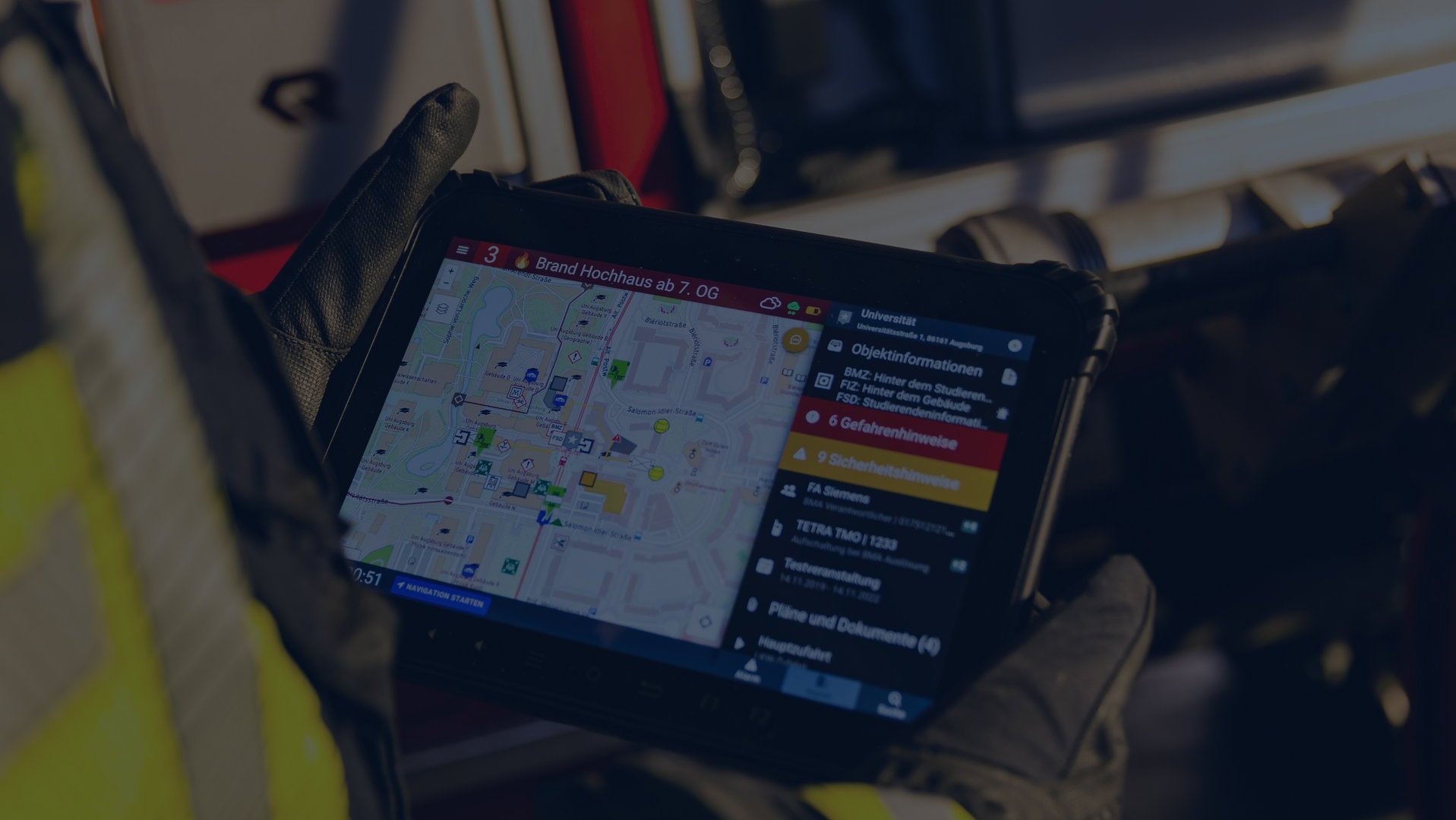 Nahaufnahme eines Einsatztablets, das von einer Einsatzkraft in Schutzkleidung gehalten wird und die Kartenübersicht für Gefahrenhinweise der Alamos Software-Lösungen zeigt. Im Hintergrund sind unscharf Einsatzklamotten mit gelben Leuchtstreifen zu erkennen. Das Bild wurde mit einer vollflächigen dunkelblauen Überlagerung abgedunkelt.