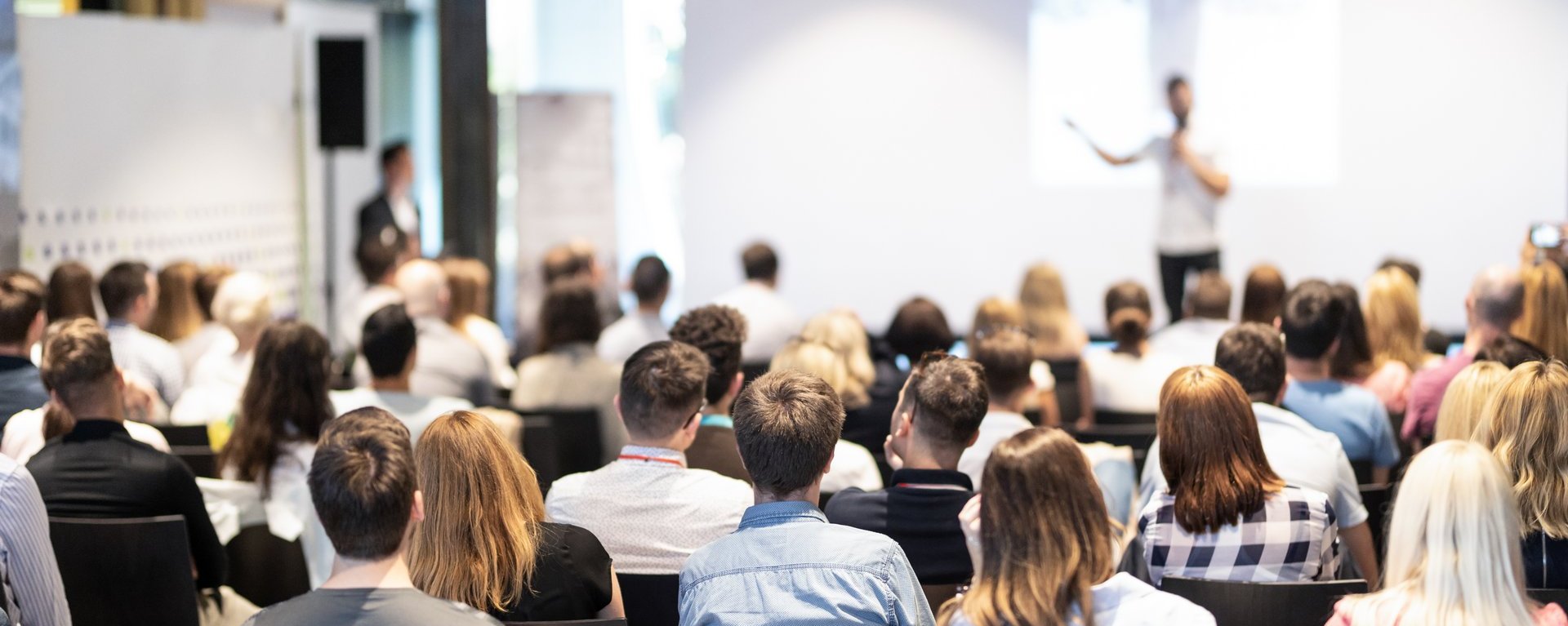 Ein Vortragender steht auf einer Bühne vor einem großen weißen Screen und hält eine Informationsveranstaltung vor einem Publikum. Das Publikum ist dabei von hinten zu sehen und steht im eigentlichen Fokus des Bildes, während der Vortragende nur unscharf in der Ferne zu erkennen ist.