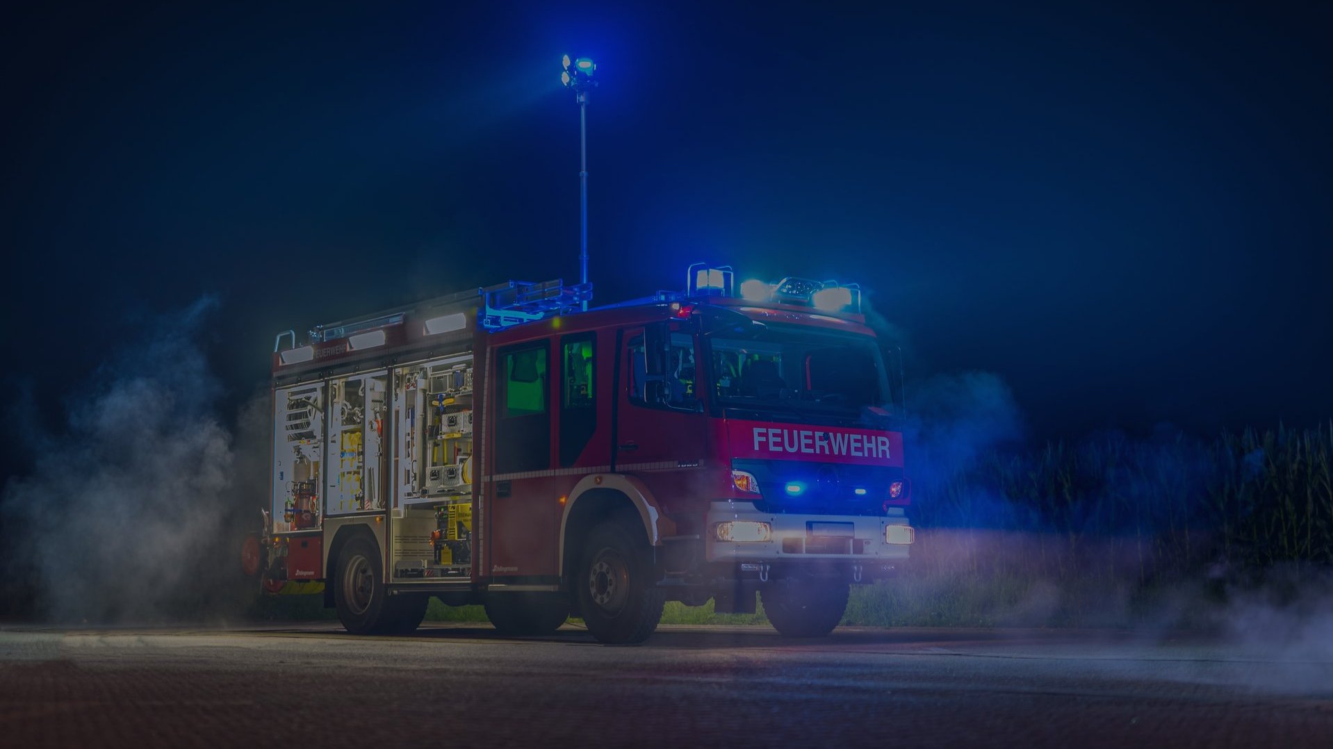 Großer Einsatzwagen der Feuerwehr mit offener Seitenklappe, in der alle möglichen Werkzeuge und einsatzrelevante Gegenstände zu sehen sind. Der Wagen steht Nachts auf einer nebligen Straße und steht in voller Beleuchtung seiner Einsatzleuchten. Das Bild wurde mit einer vollflächigen dunkelblauen Überlagerung abgedunkelt.
