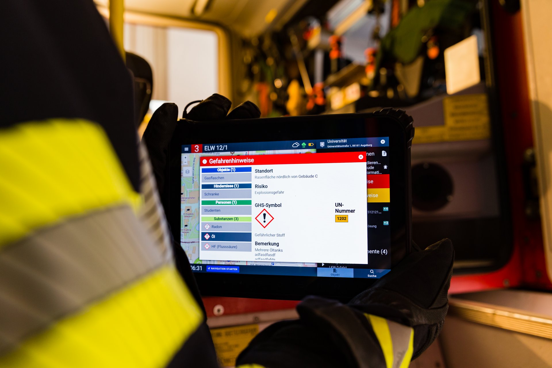 Nahaufnahme eines Einsatztablets, das von einer Einsatzkraft in Schutzkleidung in die Kamera gehalten wird. Der Screen des Tablets zeigt ein Informationsfenster mit Gefahrenhinweisen zu einem Einsatz. Im Hintergrund ist unscharf eine Werkstatt zu sehen.